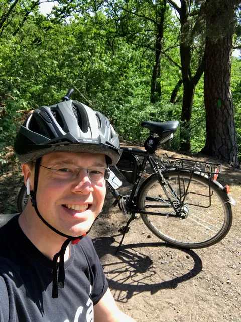 Marko Schilde, the maker of Cyclonauts, smiling in a grey cycling helmet on a forest trail, with his black touring bike and rear rack behind him